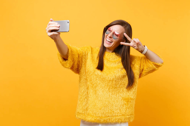 Радостная молодая женщина в меховых свитерах в очках сердца показывает знак победы делая селфи на мобильном телефоне, изолированном на ярко-желтом фоне. Люди искренние эмоции образ жизни. Рекламная зона
 - Фото, изображение