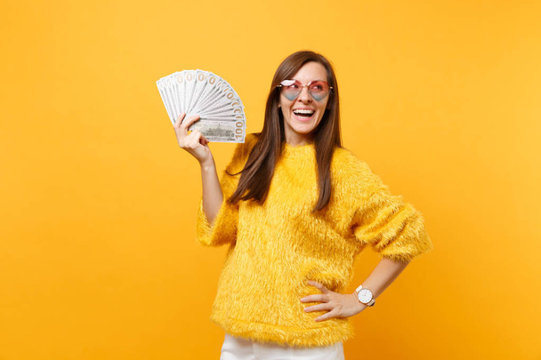 Смеющаяся молодая женщина в очках сердца смотрит вверх и держит в руках кучу долларов, наличные деньги изолированы на ярко-желтом фоне. Люди искренние эмоции, образ жизни концепции. Рекламная зона
 - Фото, изображение