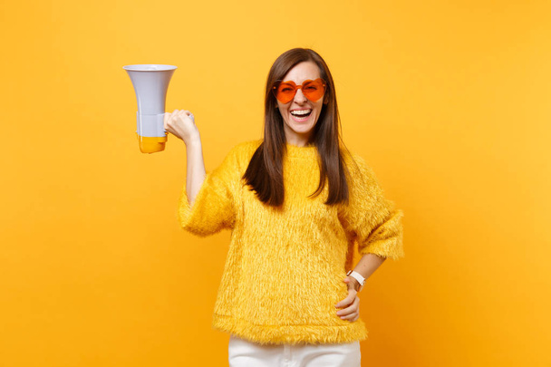 Portrait de joyeux attrayant jeune femme en chandail de fourrure orange lunettes coeur tenant mégaphone isolé sur fond jaune vif. Les gens émotions sincères, concept de style de vie. Espace publicitaire
 - Photo, image