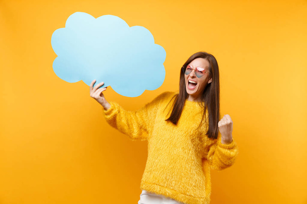 Joyful young woman in heart eyeglasses hold empty blank blue Say cloud speech bubble doing winner gesture screaming isolated on yellow background. People sincere emotions, lifestyle. Advertising area - Φωτογραφία, εικόνα