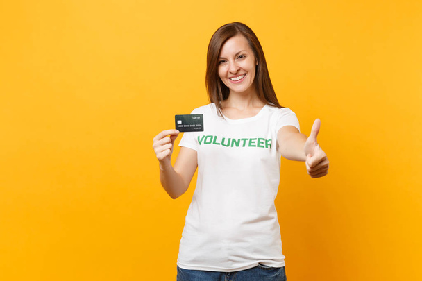 Portrét mladé ženy v bílém tričko s písemným zelený nápis titul dobrovolnická drží banky kreditku izolované na žlutém pozadí. Dobrovolnou bezplatnou pomoc pomoc, charita grace práce concept - Fotografie, Obrázek