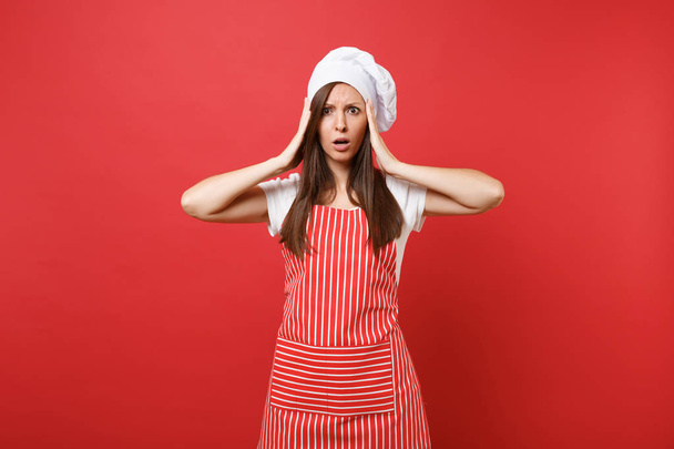 Νοικοκυρά θηλυκό σεφ μάγειρας ή φούρναρης με ριγέ ποδιά, λευκό t-shirt, Τόκα σεφ καπέλο απομονώνονται σε φόντο κόκκινο τοίχο. Λυπημένος αναστατωμένος οικονόμος σοκαρισμένος γυναίκα βάλει τα χέρια στο κεφάλι. Χλευάσουμε επάνω το αντίγραφο διαστημικής έννοιας - Φωτογραφία, εικόνα