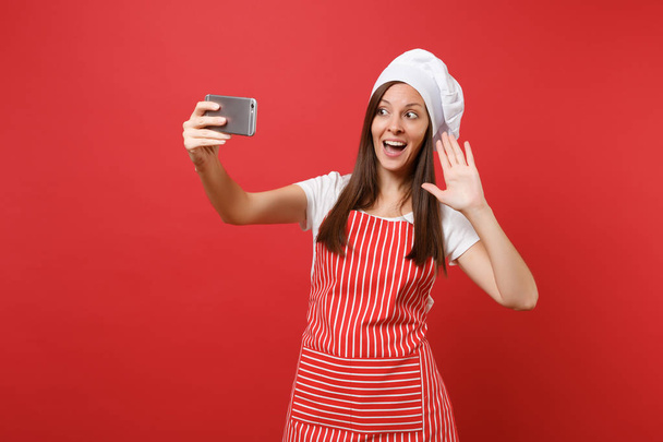Housewife female chef cook or baker in striped apron, white t-shirt, toque chefs hat isolated on red wall background. Smiling fun woman doing selfie shot on mobile phone. Mock up copy space concept - Photo, Image