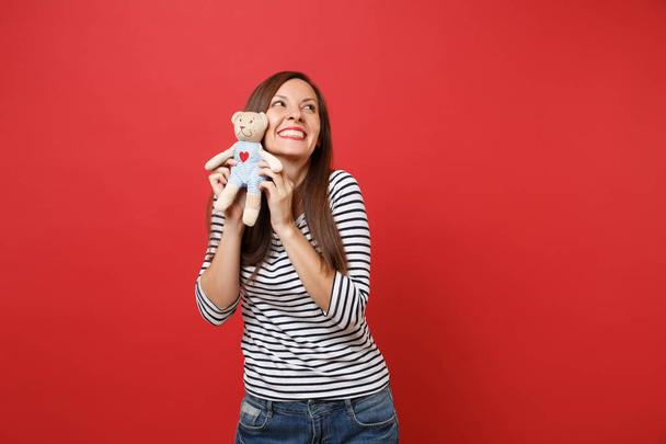 Portrait of dreamy smiling young woman in striped clothes looking up holding teddy bear plush toy isolated on bright red wall background. People sincere emotions lifestyle concept. Mock up copy space - Φωτογραφία, εικόνα