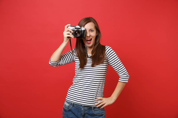 Portrait of cheerful young woman in striped clothes taking picture on retro vintage photo camera isolated on bright red wall background. People sincere emotions, lifestyle concept. Mock up copy space - Photo, image