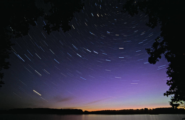 Длинные звездные следы, вращающиеся вокруг Полярной звезды. Aurora Borealis светится зеленым и фиолетовым над горизонтом
. - Фото, изображение