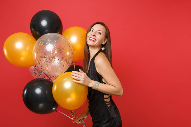 Радостная молодая женщина в маленьком черном платье празднует проведение воздушных шаров изолированы на красном фоне. St. Valentine 's, International Women' s Day, Happy New Year, birthday mockup holiday party concept
 - Фото, изображение