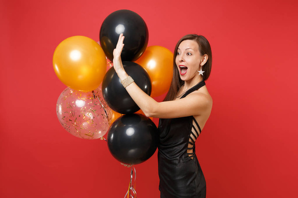 Amazed young woman in black dress celebrating, waving hand for greeting holding air balloons isolated on red background. International Women's Day Happy New Year birthday mockup holiday party concept - Photo, Image