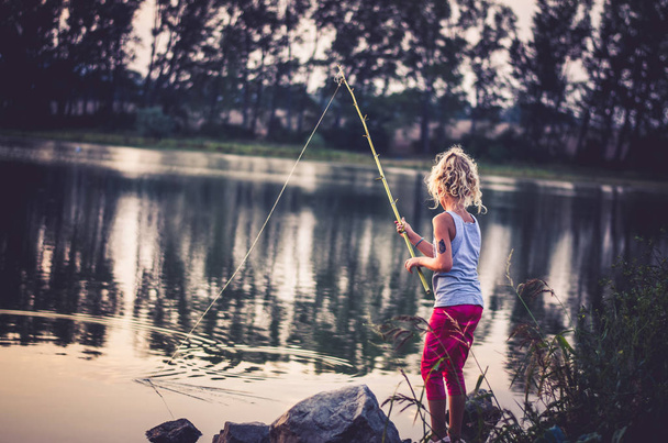schattig klein meisje vissen met zelfgemaakte hengel bij de vijver bij de zonsondergang - Foto, afbeelding
