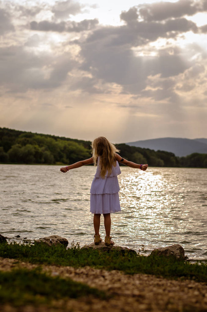 αξιολάτρευτο κοριτσάκι στο φόρεμα σε λίμνη στην ύπαιθρο το απόγευμα χαλαρώνοντας στο ηλιοβασίλεμα πίσω  - Φωτογραφία, εικόνα