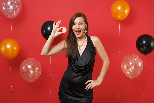 Mujer joven emocionada en vestido negro celebrando la celebración de moneda de metal bitcoin de color dorado, moneda futura en globos de aire de fondo rojo brillante. Feliz Año Nuevo, concepto de fiesta de cumpleaños simulada
 - Foto, Imagen