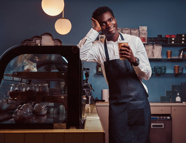 Barista Africano sorrindo para a câmera relaxante após o dia de trabalho com café, enquanto se inclina no balcão no café
. - Foto, Imagem