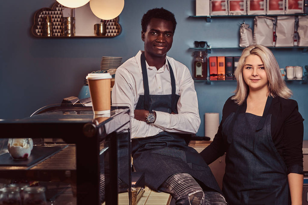 Two young multiracial baristas in aprons standing welcomingly at their trendy coffee shop. - Photo, Image