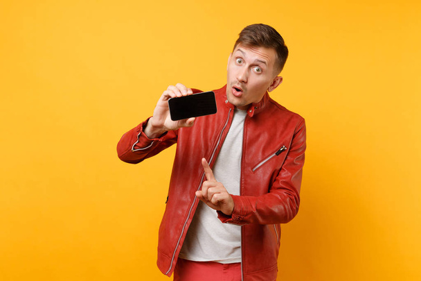 Portrait vogue amusant jeune homme en veste en cuir rouge, t-shirt tenir téléphone portable avec écran vide vierge isolé sur fond jaune vif. Les gens sincères émotions concept de style de vie. Espace publicitaire
 - Photo, image