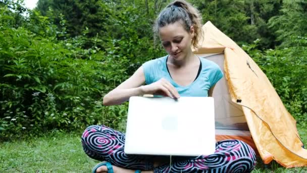 Mujer turista freelancer abre un portátil en un camping y trabaja en la naturaleza sobre el telón de fondo de una tienda de campaña turística
. - Metraje, vídeo