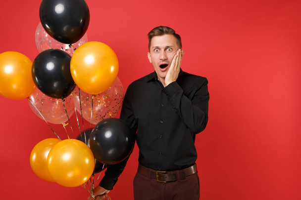 Šokovaný mladý muž v černé klasické tričko drží balónů, držet ruku na tvář izolovaných na jasně červeném pozadí. Mezinárodní den žen, šťastný nový rok narozeniny maketa holiday party koncept - Fotografie, Obrázek