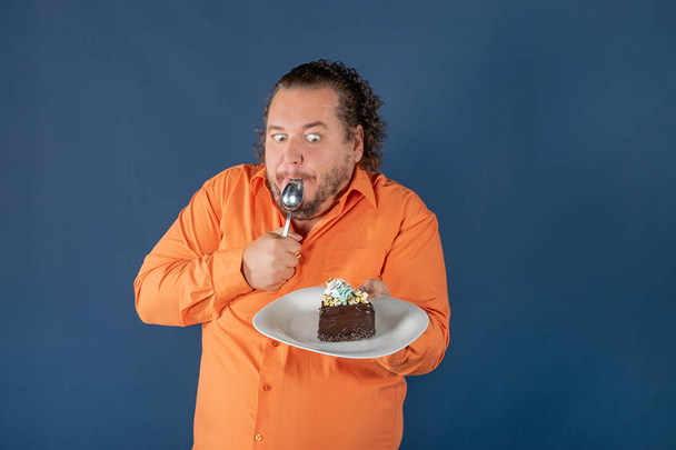 Funny fat man in orange shirt with a piece of chocolate cake on a plate. Birthday celebration - Photo, image