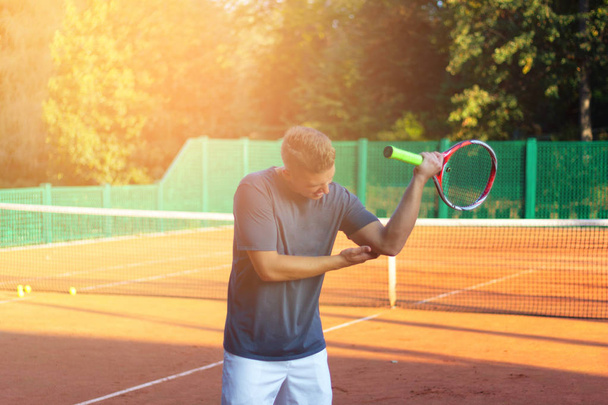 Handsome man on tennis court. Young tennis player. Pain in the elbow with sunlight in background - Фото, зображення