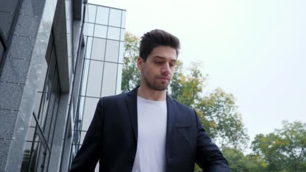 Young man in hurry late for work. Handsome businessman or student looks at watch and with quick walking to the meeting. Male model on office building background. - Imágenes, Vídeo