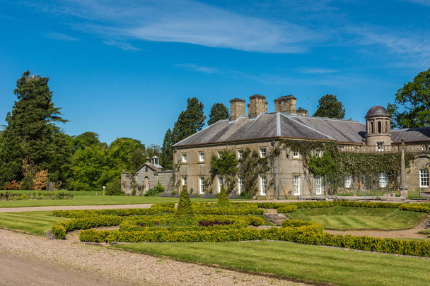 Камнок, Айршир, Шотландия, Великобритания - 18 июня 2012 года: вид сбоку на каменную секцию дома Дамфрис под голубым небом, зеленый сад спереди. Темно-зеленые деревья в спину
. - Фото, изображение
