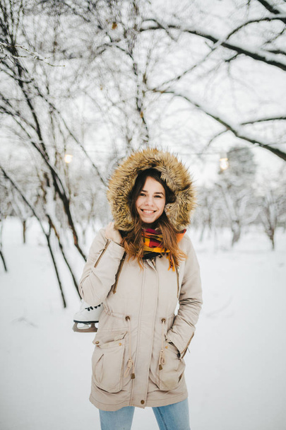A beautiful young Caucasian woman standing in a snow-covered park in a jacket with a hood and fur on her head in jeans and holding on her shoulder a pair of white skates for long shoelaces. - Photo, Image