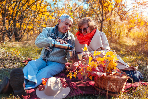 Starší pár s čajem v podzimním lese. Šťastný muž a žena užívat piknik a přírodou. Usmívající se muž vylévající čaj z termosky - Fotografie, Obrázek