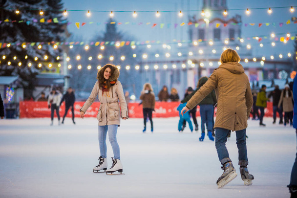 Young couple in love Caucasian man with blond hair with long hair and beard and beautiful woman have fun, active date ice skating on the ice arena in the evening city square in winter on Christmas Eve - Фото, зображення