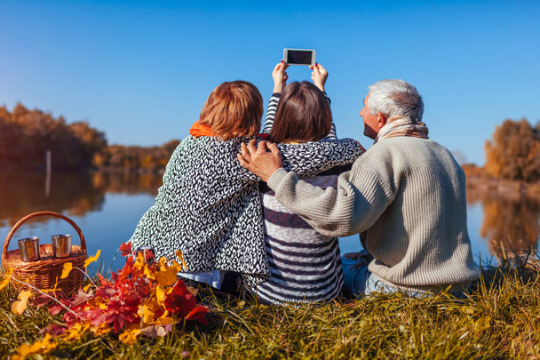 I genitori anziani prendono selfie al telefono dal lago autunnale con la loro figlia adulta. Valori familiari. Persone che fanno picnic
 - Foto, immagini