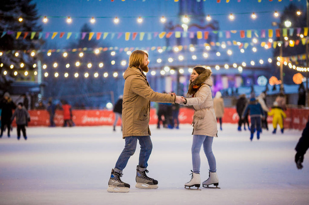 Uzun saçlı sarışın saçlı beyaz adam genç çift tr ve eğlenceli, aktif tarihi buz pateni Buz arenada akşam şehir meydanında Noel arifesinde kışın sakal ve güzel bir kadın var - Fotoğraf, Görsel