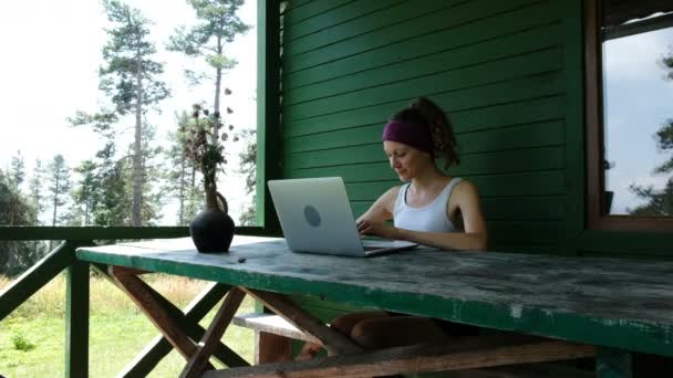 Χαμογελώντας επιχειρήσεων γυναίκα τουριστικά ελεύθερος επαγγελματίας πληκτρολογώντας σε ένα φορητό υπολογιστή σε εξωτερικούς χώρους το καλοκαίρι ενάντια σε έναν πράσινο τοίχο σε ένα κέντρο αναψυχής μέσα στο δάσος - Πλάνα, βίντεο