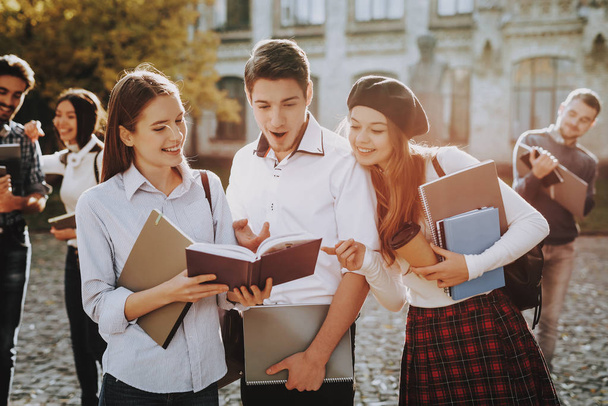Holding Books. Two Girls and Boy. Knowledge. Standing in University. Together. Courtyard. Sunny Day. Students. Good Mood. Intelligence. Standing Together. Outside. Friends. Happy. Books. University. - 写真・画像