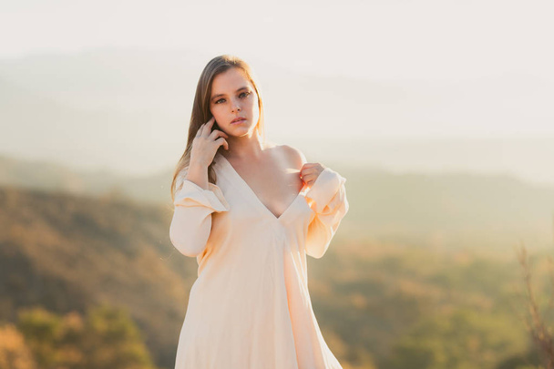 Young mixed race woman in white dress standing above a valley at sunrise - Photo, Image