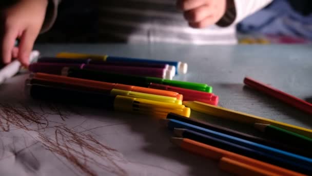 Pequeño niño elige plumas y lápices de fieltro de colores en la mesa en casa en un pedazo de papel en la oscuridad
 - Metraje, vídeo