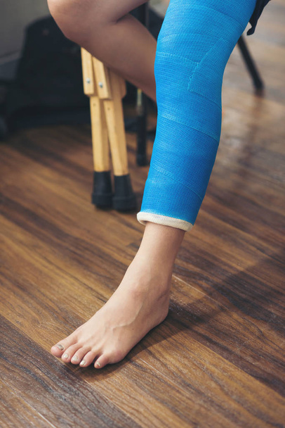 injury woman wearing black dress with blue leg splint sitting on chair and holding wooden crutches near window at hospital - Φωτογραφία, εικόνα