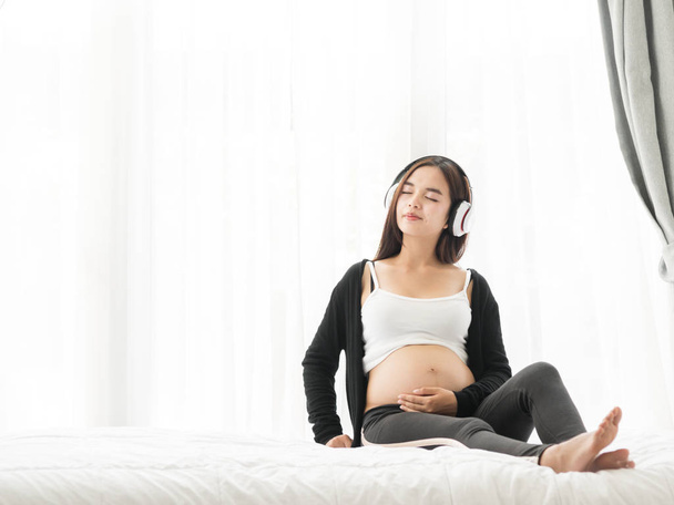 Beautiful Asian pregnant woman using headphone on her belly at home, lifestyle concept. - Foto, immagini