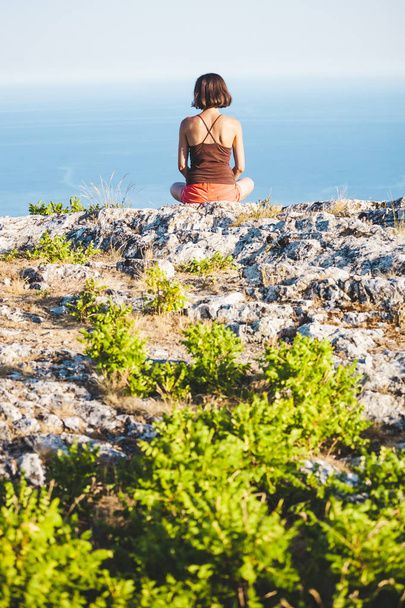 Ein Mädchen sitzt bei Sonnenuntergang auf einem Berg. eine Frau macht Yoga und sitzt in einer Lotusposition. Meditation ist weit von Zivilisation entfernt. schöner weiblicher Rücken. Blick auf die Berge und das Meer. - Foto, Bild
