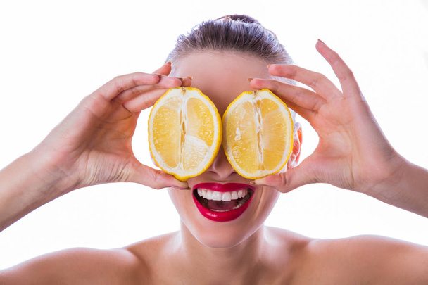 Close-up young joyful woman with bare shoulders keeps two halves of lemon in hands which hides her eyes, on the white background - Foto, Imagen