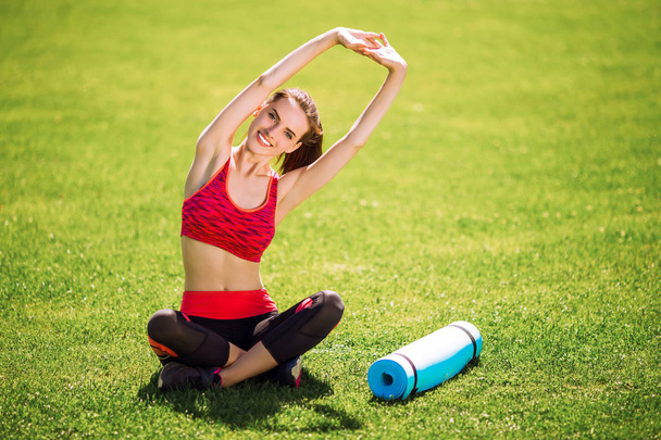 Sporty cheerful woman in sport dress sits on grass and does stretching exercises - Фото, зображення