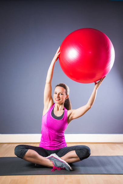 Young sporty cheerful woman in sport dress sits on carpet and keeps fitball in hands indoors - Photo, Image