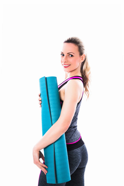 Cheerful sporty woman stands in sport dress and keeps blue carpet against the white background - Foto, afbeelding