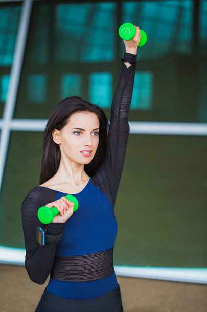 Cheerful sporty woman stands with the dumbbells in hands and shakes arms, outdoors - Foto, immagini