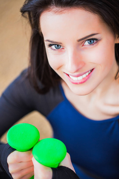Close-up face of happy sporty woman who smiles and keeps the green dumbbells in hands, outdoors - Photo, image