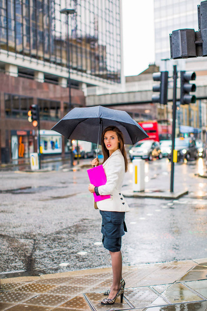 Nuori tyylikäs liiketoiminnan nainen seisoo jalkakäytävällä kadulla ja pitää kansion ja avasi sateenvarjon käsissä
 - Valokuva, kuva