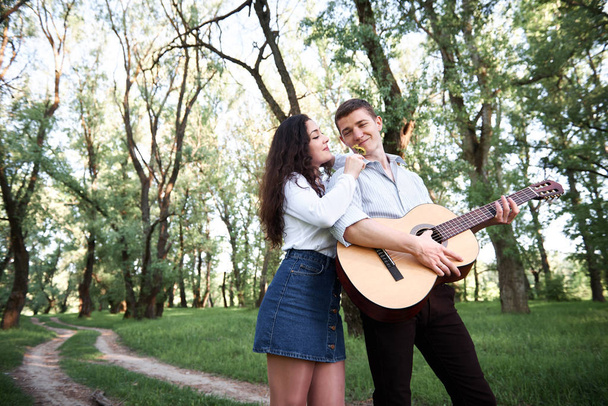 young couple walking in the forest and playing guitar, summer nature, bright sunlight, shadows and green leaves, romantic feelings - Zdjęcie, obraz