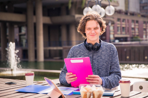 Handsome smiling guy sits with the tablet, with cup of coffee,with food, with folders and he keeps one folder in his hands in time of the break, outdoor - Fotoğraf, Görsel
