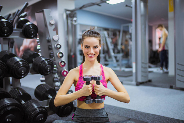 Young sports woman sits on the floor and keeps dumbbells in hands, at the gym - Фото, зображення