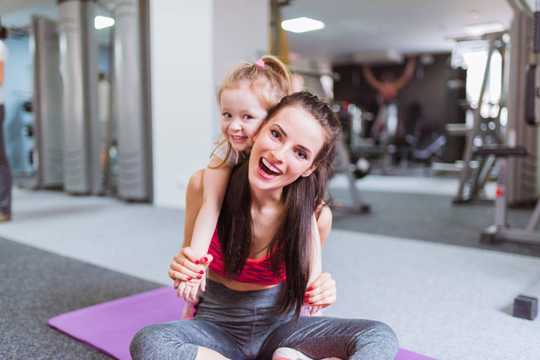 Happy young mother sits on the carpet and keeps on her back her daughter, in the gym - Foto, Imagen