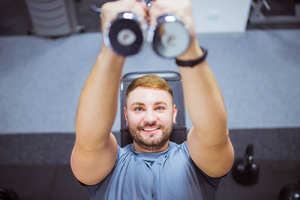 Primer plano feliz entrenador muscular se encuentra en un simulador y hace ejercicios con las mancuernas en sus manos, en el gimnasio
 - Foto, Imagen