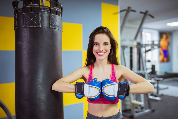 Happy smiling sports woman stands near punching bag dressed in boxing glove, at the gym - 写真・画像