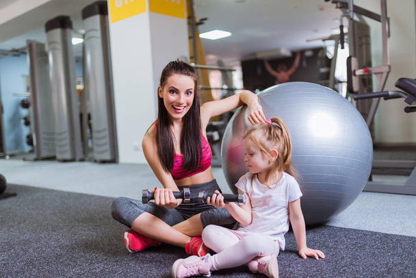 Beautiful young mother sits together with her daughter on the floor near fitball and they keep dumbbells in their hands, at the gym - Photo, Image